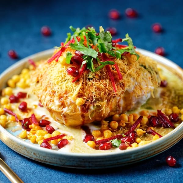A vibrant Indian chaat dish, topped with sev, pomegranate, cilantro, and sauces on a blue background.