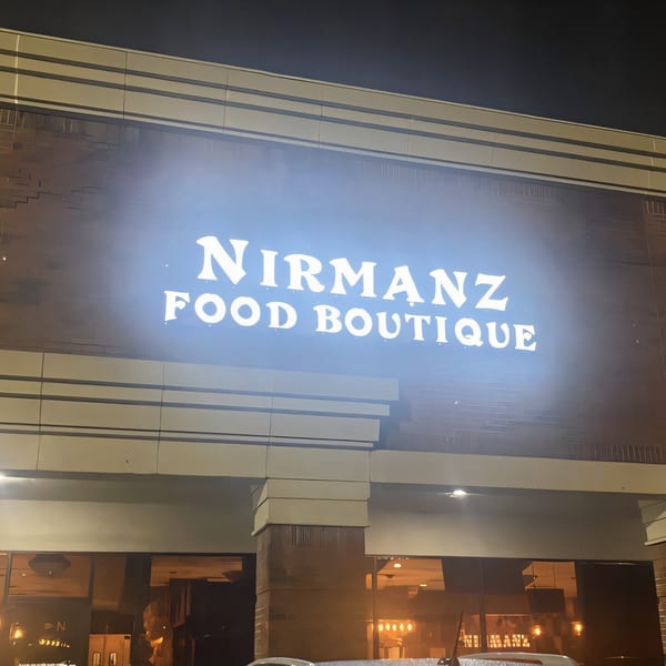The illuminated "Nirmanz Food Boutique" sign glows brightly on the restaurant's brick exterior at night.