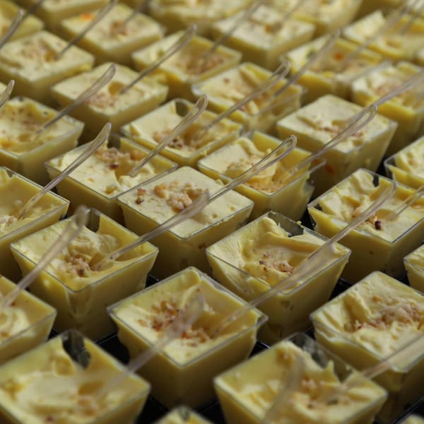 Rows of small square cups filled with yellow Indian dessert, topped with nuts and tiny spoons.