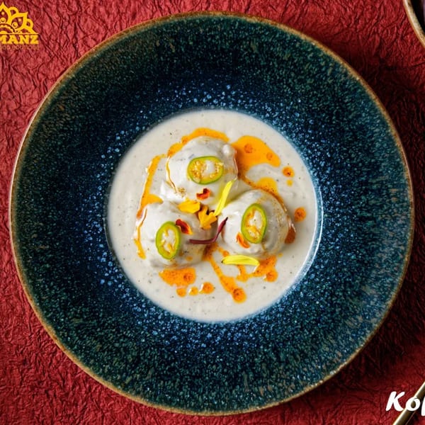 Three Indian kofta dumplings in creamy white sauce with chili and yellow petals on a blue plate.