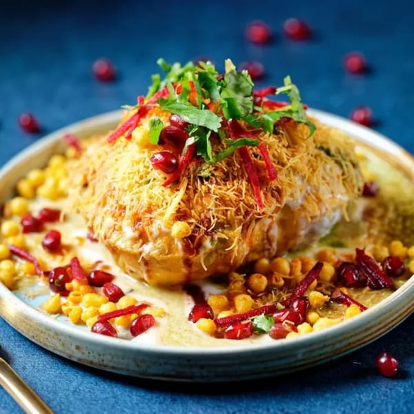 A vibrant Indian chaat dish, topped with sev, cilantro, pomegranate seeds, and sauces on a blue background.