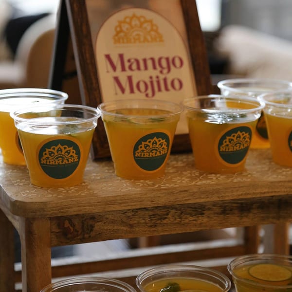 Nirmanz Food Boutique's Mango Mojito samples in plastic cups on a wooden table with a sign.