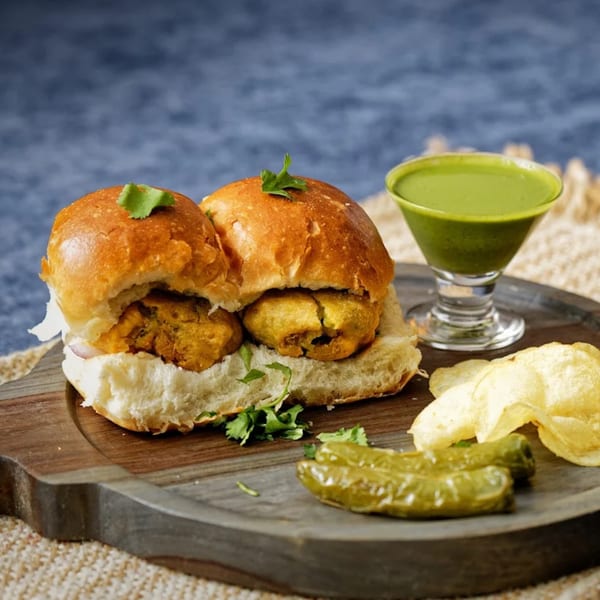 Two Nirmanz Food Boutique vada pavs with green chutney, fried chilies, and potato chips.